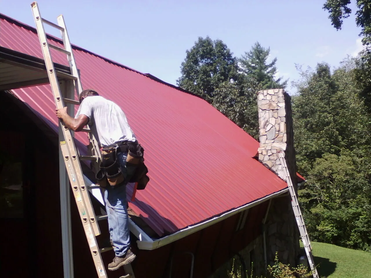 Roofer on a ladder beside a red metal roof and stone chimney — Soffit & Fascia Repair by Roofing Brothers in East Fultonham